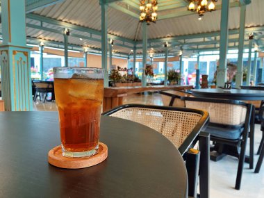 Koyu, yuvarlak bir masada ön planda belirgin bir şekilde duran ferahlatıcı bir bardak buzlu çay. Çay önü açık bir bardakta sunuluyor. Tahta bir bardakta dinleniyor. Üstünde görünen buz küpleri yüzüyor..