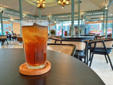 Koyu, yuvarlak bir masada ön planda belirgin bir şekilde duran ferahlatıcı bir bardak buzlu çay. Çay önü açık bir bardakta sunuluyor. Tahta bir bardakta dinleniyor. Üstünde görünen buz küpleri yüzüyor..