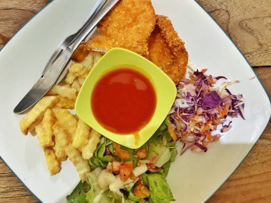 Canlı ve dengeli bir yemek, beyaz kare bir tabakta servis edilir, ince mavi kenarlı, kırsal bir ahşap masada dinlenir. Tavuk şnitzeli ya da balık ve patates kızartması tabağına benziyor. İki altın-kahverengi fileto ile karakterize edilmiş..