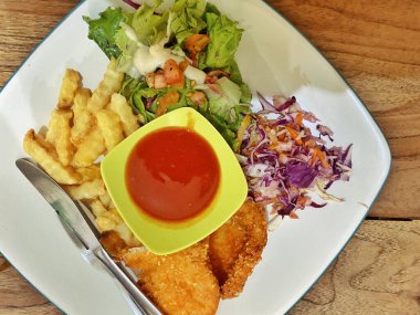 Canlı ve dengeli bir yemek, beyaz kare bir tabakta servis edilir, ince mavi kenarlı, kırsal bir ahşap masada dinlenir. Tavuk şnitzeli ya da balık ve patates kızartması tabağına benziyor. İki altın-kahverengi fileto ile karakterize edilmiş..