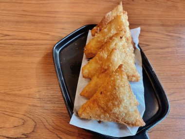 Bir tabak mükemmel altın, çıtır Samosas (veya kızartılmış wonton / empanada), modern siyah dikdörtgen tabakta servis edilir, ahşap saplı. Üçgenlerin kabuğunda çok güzel kabarcıklı bir doku var..