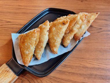 Bir tabak mükemmel altın, çıtır Samosas (veya kızartılmış wonton / empanada), modern siyah dikdörtgen tabakta servis edilir, ahşap saplı. Üçgenlerin kabuğunda çok güzel kabarcıklı bir doku var..
