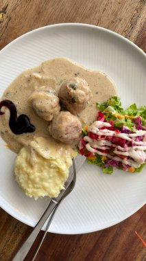 Beyaz desenli tabakta servis edilen İsveç usulü köfteler. Üç büyük, baharatlı köfte yumuşak, açık kahverengimsi bir sosta bol bol boğulur..