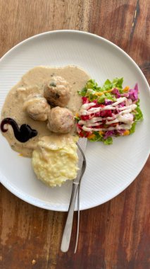 Beyaz desenli tabakta servis edilen İsveç usulü köfteler. Üç büyük, baharatlı köfte yumuşak, açık kahverengimsi bir sosta bol bol boğulur..