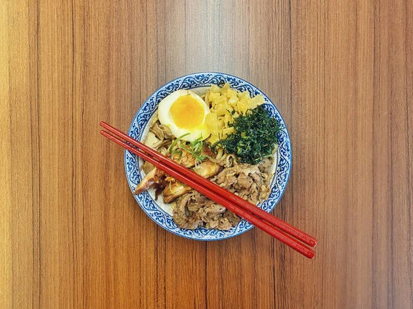 Mavi ve beyaz desenli seramik bir kasede servis edilen canlı bir Japon pirinç kasesinin yukarıdan aşağı görüntüsü. Bir çift parlak kırmızı çubuk üst tarafta köşegen olarak duruyor..