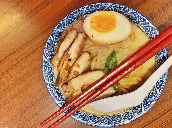 Ilık ahşap bir masanın üstünde duran mavi ve beyaz desenli seramik kasede titizlikle düzenlenmiş klasik bir ramen kasesinin canlı, tepeden tırnağa görüntüsü..