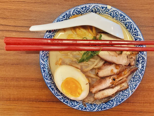 Ilık ahşap bir masanın üstünde duran mavi ve beyaz desenli seramik kasede titizlikle düzenlenmiş klasik bir ramen kasesinin canlı, tepeden tırnağa görüntüsü..