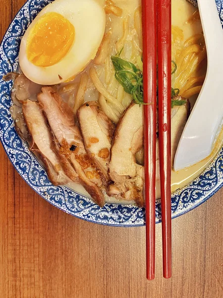 Ilık ahşap bir masanın üstünde duran mavi ve beyaz desenli seramik kasede titizlikle düzenlenmiş klasik bir ramen kasesinin canlı, tepeden tırnağa görüntüsü..