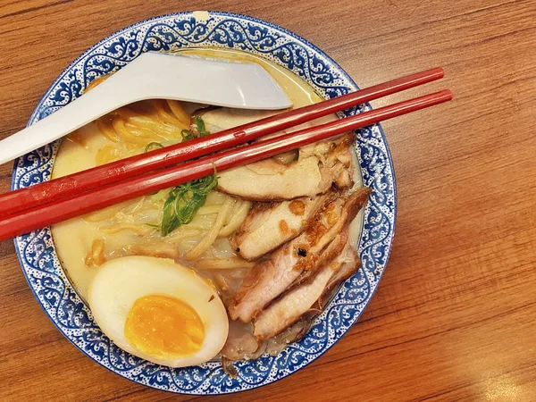 Ilık ahşap bir masanın üstünde duran mavi ve beyaz desenli seramik kasede titizlikle düzenlenmiş klasik bir ramen kasesinin canlı, tepeden tırnağa görüntüsü..