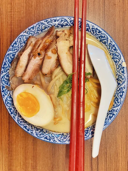 Ilık ahşap bir masanın üstünde duran mavi ve beyaz desenli seramik kasede titizlikle düzenlenmiş klasik bir ramen kasesinin canlı, tepeden tırnağa görüntüsü..