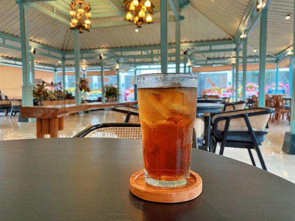 Koyu, yuvarlak bir masada ön planda belirgin bir şekilde duran ferahlatıcı bir bardak buzlu çay. Çay önü açık bir bardakta sunuluyor. Tahta bir bardakta dinleniyor. Üstünde görünen buz küpleri yüzüyor..