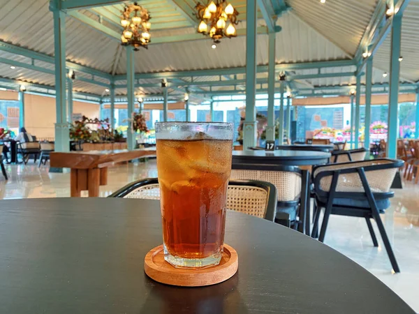 Koyu, yuvarlak bir masada ön planda belirgin bir şekilde duran ferahlatıcı bir bardak buzlu çay. Çay önü açık bir bardakta sunuluyor. Tahta bir bardakta dinleniyor. Üstünde görünen buz küpleri yüzüyor..