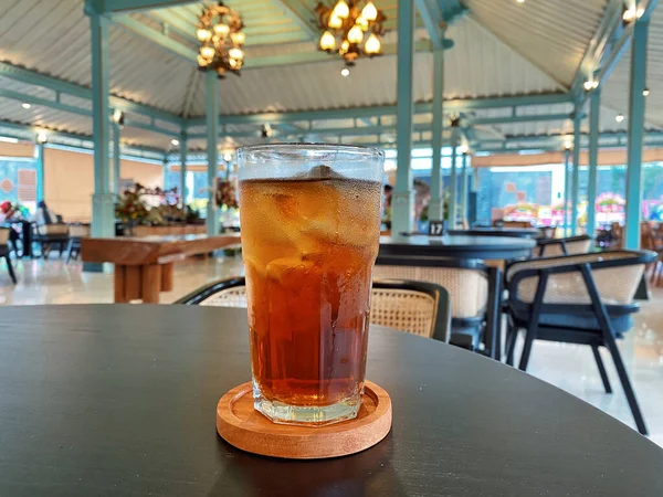 Koyu, yuvarlak bir masada ön planda belirgin bir şekilde duran ferahlatıcı bir bardak buzlu çay. Çay önü açık bir bardakta sunuluyor. Tahta bir bardakta dinleniyor. Üstünde görünen buz küpleri yüzüyor..