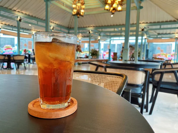 Koyu, yuvarlak bir masada ön planda belirgin bir şekilde duran ferahlatıcı bir bardak buzlu çay. Çay önü açık bir bardakta sunuluyor. Tahta bir bardakta dinleniyor. Üstünde görünen buz küpleri yüzüyor..