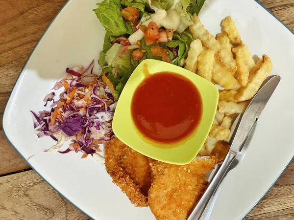 Canlı ve dengeli bir yemek, beyaz kare bir tabakta servis edilir, ince mavi kenarlı, kırsal bir ahşap masada dinlenir. Tavuk şnitzeli ya da balık ve patates kızartması tabağına benziyor. İki altın-kahverengi fileto ile karakterize edilmiş..