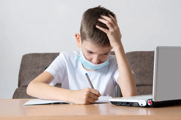 एक स्मार्ट स्कूल लड़का लैपटॉप के सामने अपने डेस्क पर बैठे हुए अपनी नोटबुक में जानकारी लिखता है। ऑनलाइन शिक्षा — स्टॉक फ़ोटो, इमेज