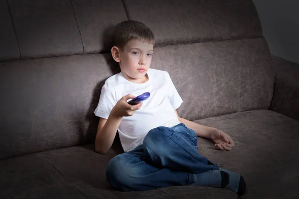 Un chico guapo está sentado en el sofá usando el mando a distancia, el niño está viendo la televisión — Foto de Stock