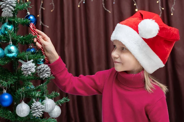 一个身穿红色毛衣 头戴圣诞帽的可爱的白人金发女孩在装饰过的圣诞树上挂着圣诞糖果 圣诞及新年概念 免版税图库图片