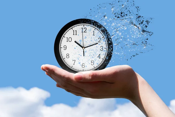 End of time or flight of time concept. A woman\'s hand holds a round clock with arrows indicating the time of 10 minutes past ten, the clock falls into small pieces against a blue sky, front view