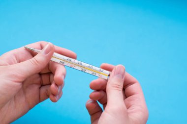 Yakın plan kadın eli, mavi arka planda bir termometre tutuyor. Termometre vücut ısısının yüksek olduğunu gösteriyor..