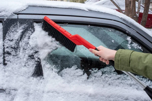 Winter problems of car drivers. Close-up of a woman\'s hand brushing snow from her car with a red brush on a cold snowy winter day.