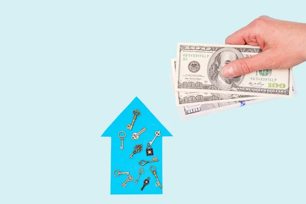 The concept of a new home, mortgage, loan. Dream home concept. Woman\'s hand holds dollars over a model blue paper house and keys on a pastel background, mock up, copy space, top view