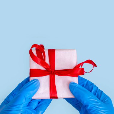 Coronavirüs salgını sırasındaki bayramınız için tebrikler. Mavi eldivenli eller mavi arka planda kırmızı kurdeleli beyaz hediye kutusu tutuyor, kare, kopyalama alanı. Sevgi gönderiyorum.