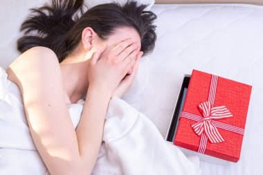 Sürpriz doğum günü konsepti. Yatakta yatan kız elleriyle gözlerini kapatıyor, önünde evde kırmızı bir hediye kutusu yatıyor. Düğün, Noel, Sevgililer Günü için kullanılabilir.