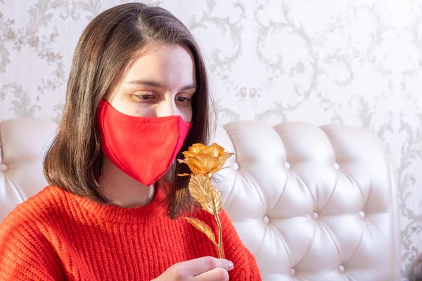 Young caucasian girl wearing a red protective mask holding a golden rose at home, front view, copy space. Can be used as postcard for Valentines Day 2021, Mothers Day, Birthday