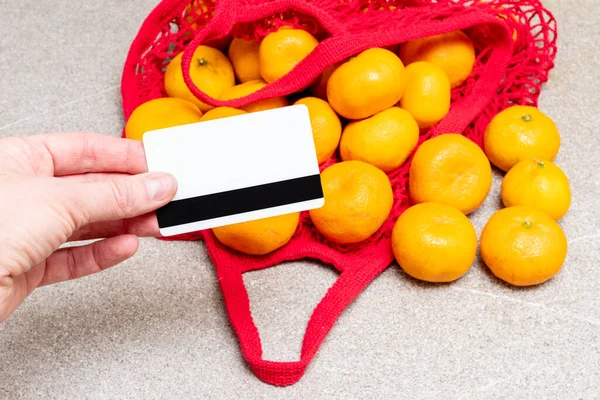 Κόκκινη τσάντα με πορτοκαλί μανταρίνια και το χέρι δίνοντας μια τραπεζική κάρτα για να πληρώσει για μια αγορά σε γκρι φόντο — Φωτογραφία Αρχείου