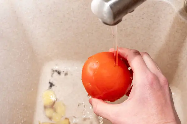 Hand hält eine rote Tomate unter fließendem Wasser in der Spüle, Draufsicht, Großaufnahme. Kochen zu Hause — Stockfoto