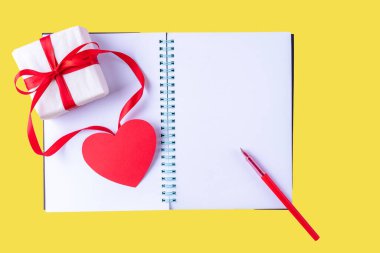 Beyaz boş defter, kırmızı kalem, kırmızı kurdeleli hediye kutusu ve sarı arka planda pembe kağıt kalp şekli.