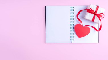 Beyaz boş not defteri, kırmızı kurdeleli hediye kutusu ve pembe arka planda pembe kağıt kalp şekli, boşluğu kopyala