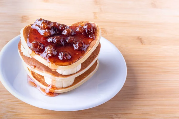 Homemade heart shaped pancakes with bananas and berry jam on a white plate on a wooden background — Stock Photo, Image