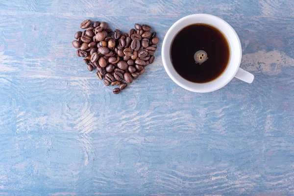 Heart shape made from coffee beans and white coffee cup on blue stylish wooden background, close-up, top view, flat lay. Symbol of love for coffee.