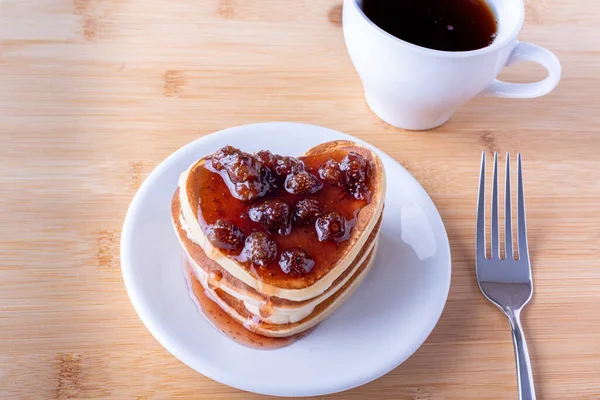 Homemade heart shaped pancakes with berry jam on white plate, fork and mug with coffee on wooden background — Stock Photo, Image