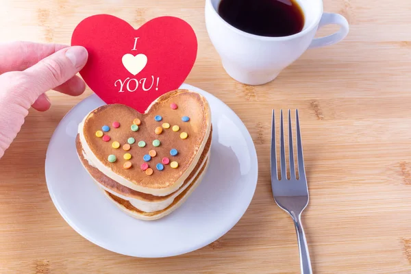 A womans hand holds a red heart with the inscription I LOVE YOU over homemade pancakes, mug with coffee, fork — Stock Photo, Image