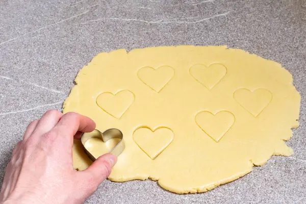 Een vrouwenhand snijdt een hartvormig koekje van geel deeg op een grijze trending keukentafel, close-up, bovenaanzicht — Stockfoto