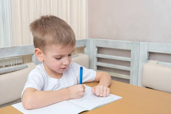 The child studies online at home. Cute little boy writes homework in a notebook sitting at the table at home. Online home education concept