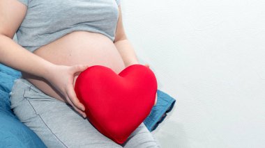 Kadın elleri, hamile bir kadının karnının arka planına karşı kalp şeklinde kırmızı bir yastık tutuyor.