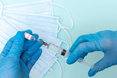 Mavi tıbbi eldivenli bir doktorun elinde mavi maskeli mavi bir arka plandaki şırıngaya aşı çözeltisi yerleştirirken. Aşı ve koronavirüs enfeksiyonu tedavisi kavramı.