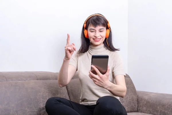 Mujer atractiva sentada en el sofá con auriculares naranjas hablando con la tableta y levantando el dedo — Foto de Stock