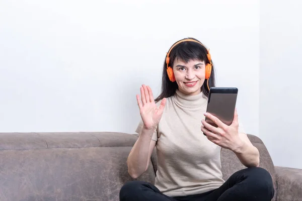 Encantadora morena con auriculares naranjas haciendo una videollamada y mostrando un gesto de hola o adiós — Foto de Stock