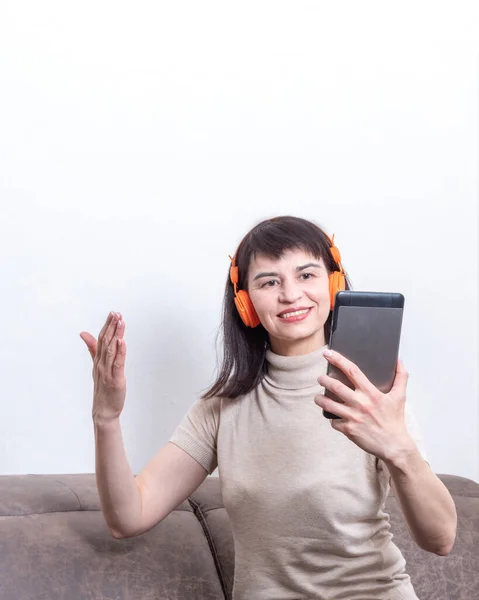 Encantadora morena sentada en un sofá con auriculares naranjas con videollamadas y gestos — Foto de Stock