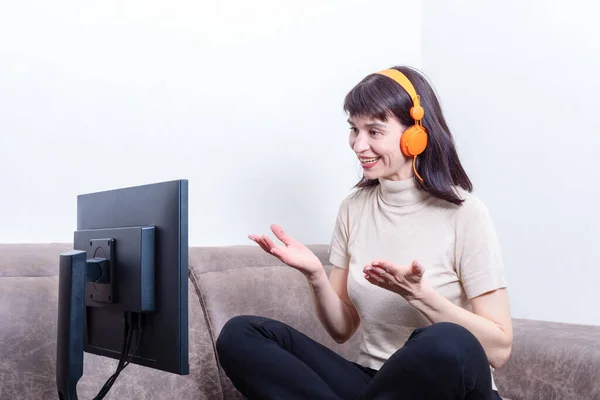 Attractive woman wearing orange headphones looking at a computer monitor, gesturing and communicating online