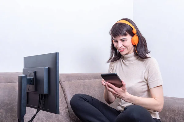 Mujer atractiva con auriculares naranjas sentada en el sofá delante de un monitor y presionando la tableta — Foto de Stock