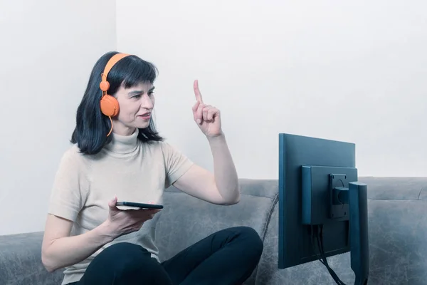 Mujer atractiva sentada con auriculares naranjas frente a un monitor de computadora y sosteniendo una tableta — Foto de Stock