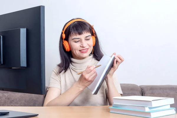 Una atractiva profesora de auriculares naranjas sentada frente a un monitor de ordenador, explica una tarea en línea — Foto de Stock