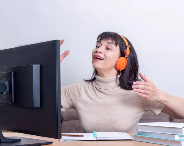 Mujer atractiva con auriculares naranjas sentados en una mesa, mirando un monitor de computadora, haciendo gestos, charlando en Internet. Concepto de aprendizaje en línea para adultos. Concepto de teletrabajo — Foto de Stock
