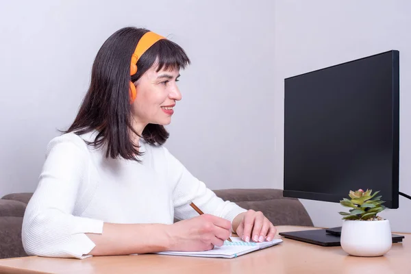Mujer sonriente con auriculares naranjas sentada en la mesa, mirando la pantalla negra en blanco del monitor, escribiendo — Foto de Stock